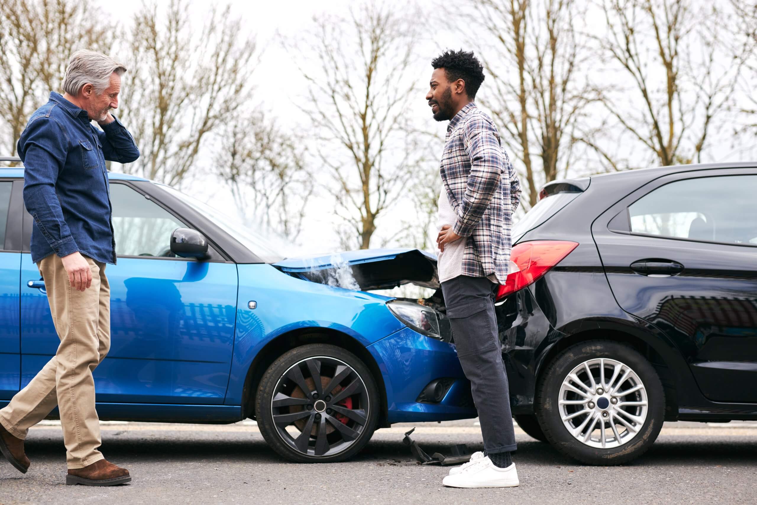 Senior and younger male drivers get out of cars and inspect damage after road traffic accident