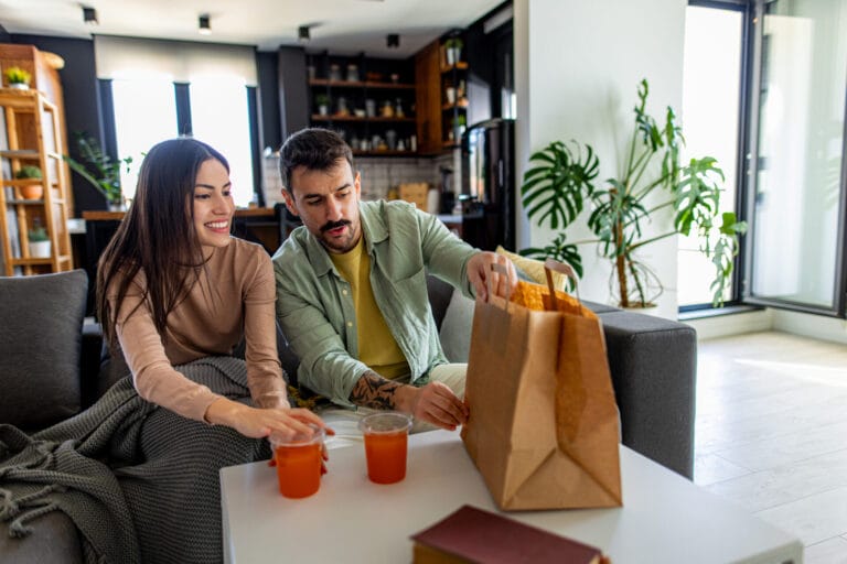 Young couple unpacking Uber eats takeaway food and enjoying a relaxing day at home, sharing delicious meals and drinks on a cozy sofa